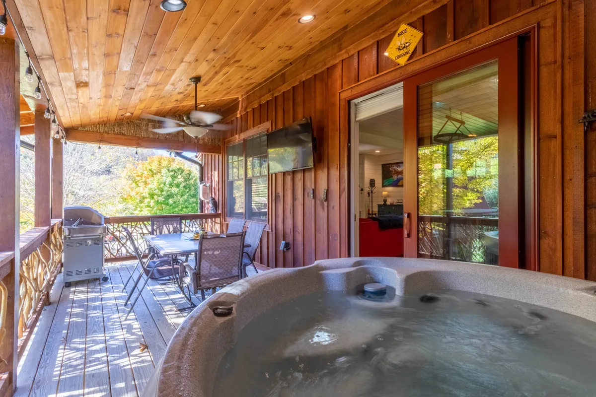 This image showcases a covered outdoor patio featuring a dining table set beneath a warm wooden ceiling. A nearby hot tub adds to the atmosphere of relaxation, and a gas grill is positioned for convenient barbecuing. The setting is cozy and inviting, with soft lighting and a view that hints at the surrounding natural beauty—perfect for outdoor dining and socializing in any weather.