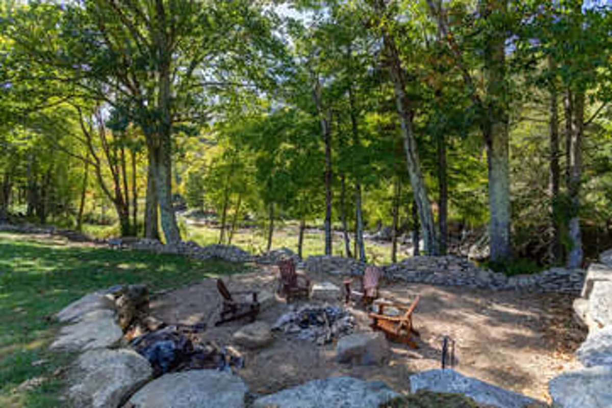 A cozy outdoor firepit area surrounded by wooden chairs and nestled among tall trees. A stone wall wraps around the space, and the forest creates a sense of seclusion. The atmosphere is perfect for couples to relax, share stories, and enjoy the tranquility of nature, especially as the sun sets or under a starlit sky.