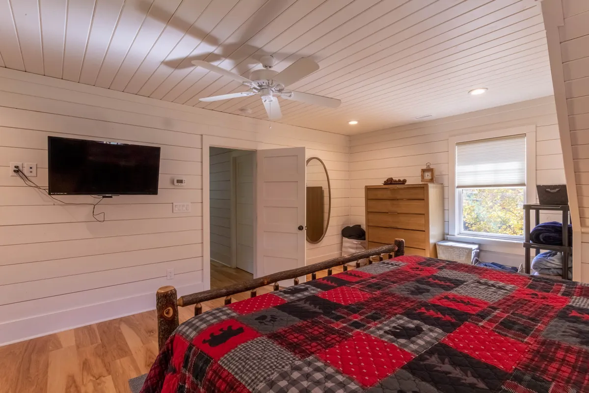 A warmly lit bedroom featuring a bed with a colorful patchwork quilt, a wooden dresser, and a window inviting in natural light. The setting exudes comfort, with subtle touches like a wall-mounted television and the gentle ambiance ideal for unwinding after a day outdoors.