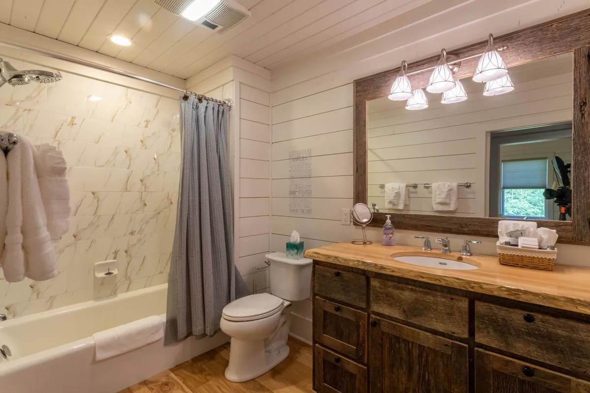 A bright and serene full bathroom with a rustic wooden vanity, large mirror with soft lighting, and a bathtub surrounded by a marble backsplash and plush towels. The space feels calm and inviting, ideal for both morning routines and soothing baths.