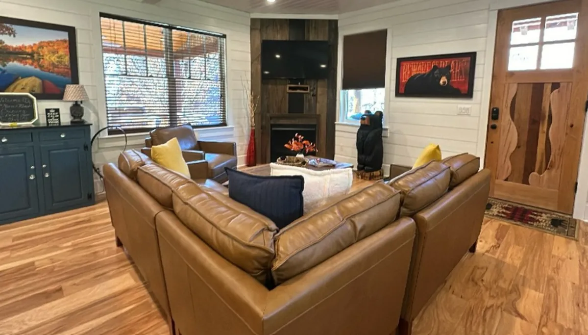 A spacious living room featuring a leather sectional sofa arranged around a cozy coffee table, large windows letting in abundant natural light, and tasteful artwork on the wall. The entrance door and the visible hardwood floors enhance the welcoming atmosphere.