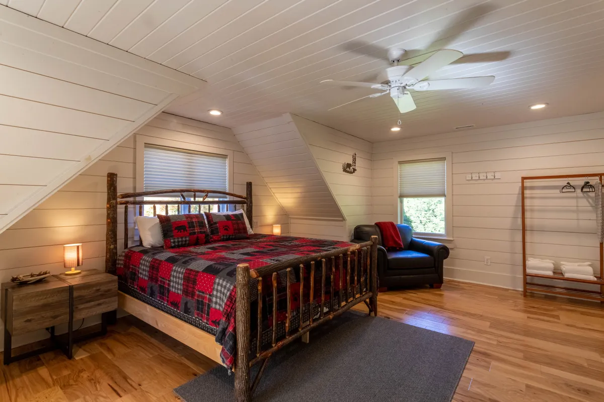 A warmly lit bedroom featuring a rustic log bed with plaid bedding, complemented by an inviting armchair and wooden flooring. Sunlight filters through the windows, creating a cozy and serene ambiance perfect for unwinding after a long day.