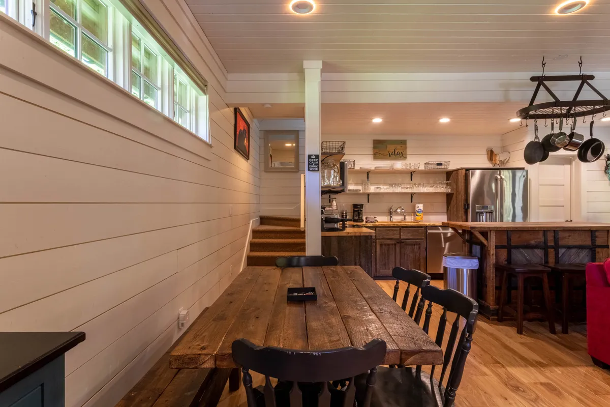 An open-concept dining and kitchen area with a wooden dining table for four, a visible kitchen area with a bar counter, and a staircase leading to the upper level. The warm lighting and wood accents provide a welcoming ambiance, tying together the dining and kitchen spaces.