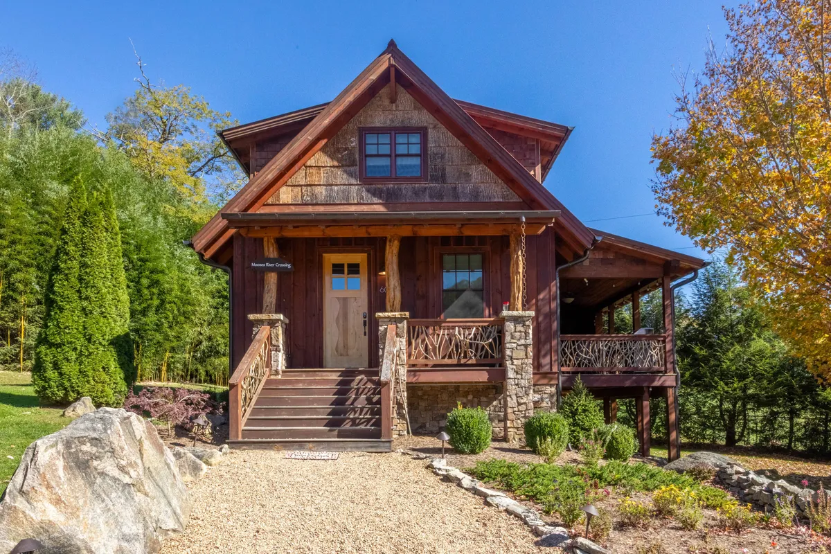 The image showcases the inviting exterior of a charming wooden cabin, with a welcoming front porch set above stone accents and wide wooden steps. Lush landscaping surrounds the cabin, creating a tranquil, nature-filled setting ideal for both relaxation and gatherings. The porch offers picturesque views and a cozy spot to unwind, while the garden hints at opportunities for outdoor activities like barbecues or morning coffee with the sounds of the nearby river.