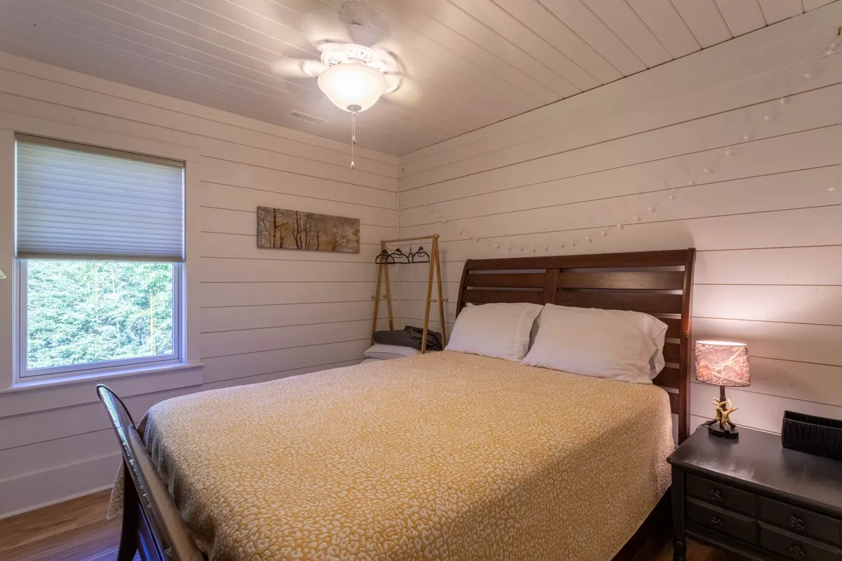 A tranquil bedroom scene with a plush bed, nightstand, soft artwork, and a window that frames views of the surrounding natural beauty. String lights add a cozy touch, and the space feels both restful and intimately connected to the outside environment.